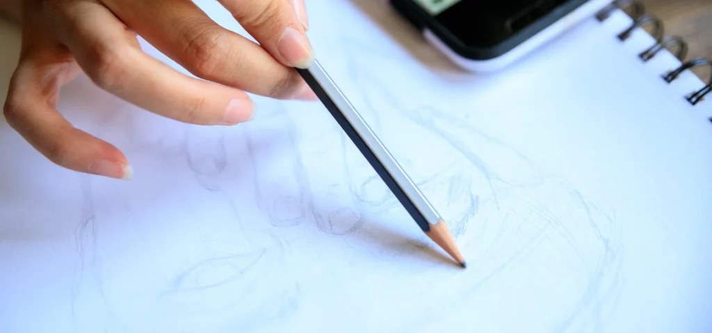 in the center of the image is a faber castell pencil that is lightly sketching on the paper. the persons hand is coming from the top left hand side of the image and they are lightly holding the pencil. the paper being drawn on is a sketch book that is ring bound as you can see the black binds on the right hand side of the image. the top right hand side is a phone where they are copying the image from.