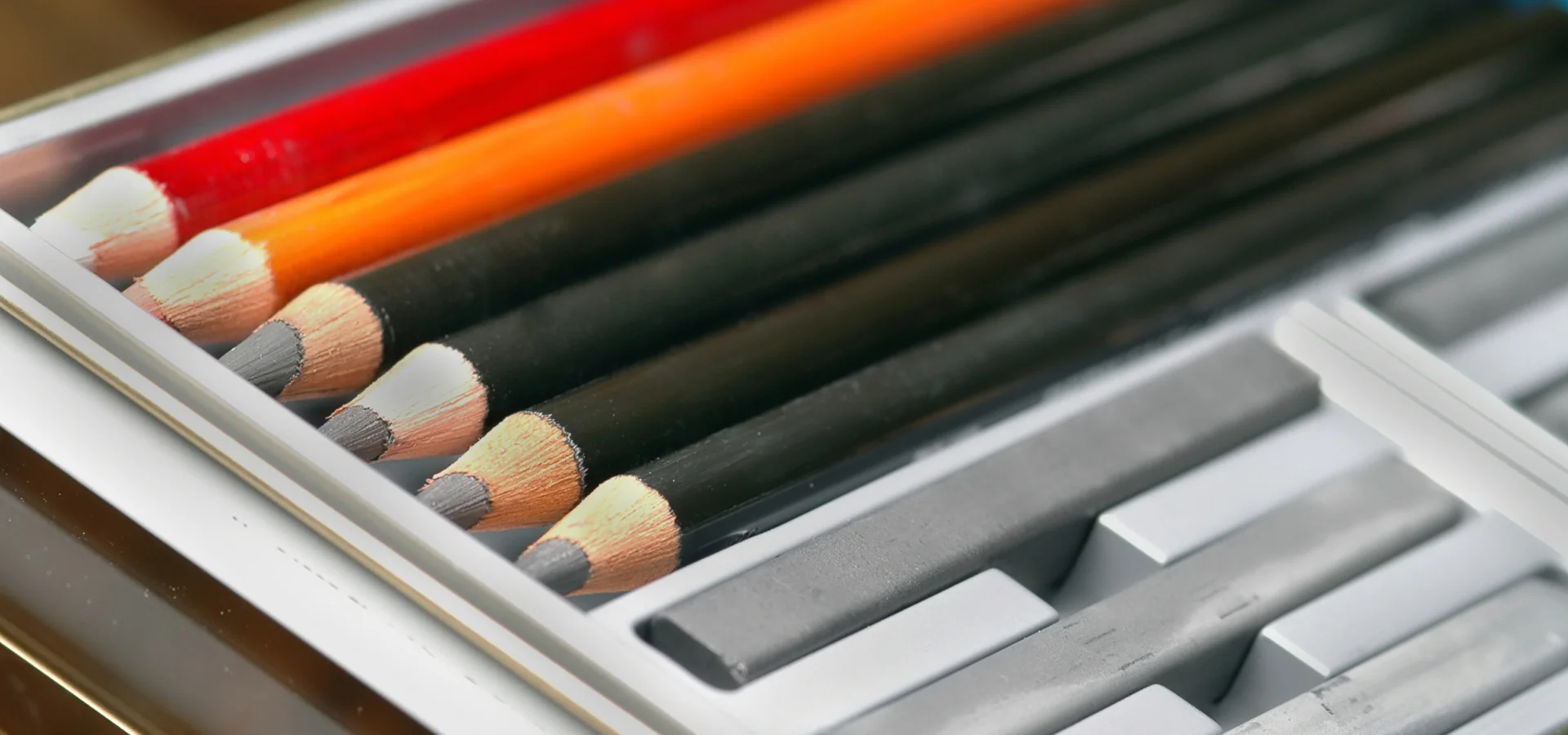 sketching-pencil-sets-a-doodle-dream-come-true shows a close up of a tin of sketching pencils and graphite blocks. the pencils on the far left are red and orange and the four closer ones are black cases. they all have grey nibs. closest to the front of the image are the sticks of compressed graphite. they are all inside of the tin. they are resting on a brown wooden table. the light is coming directly at them.