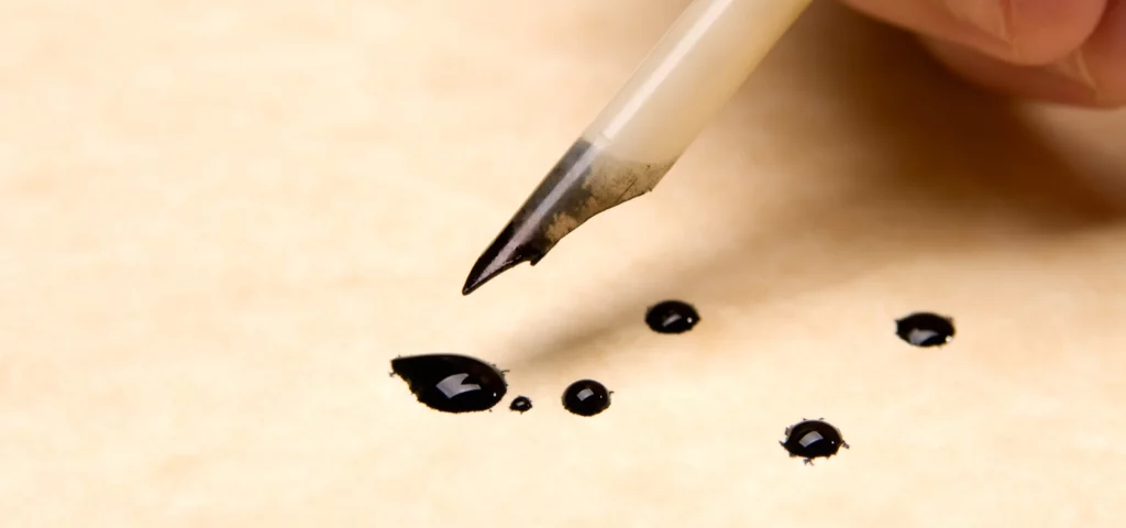 speedball acrylic on is shwon in the image. on the right hand side a hand is coming into the image. it is holding an old fashioned quill. you can oly see the top of the quill pen. there is black ink on the nib of it. below the pen is parchment paper that has black dots of ink that have been dropped onto it by the pen.