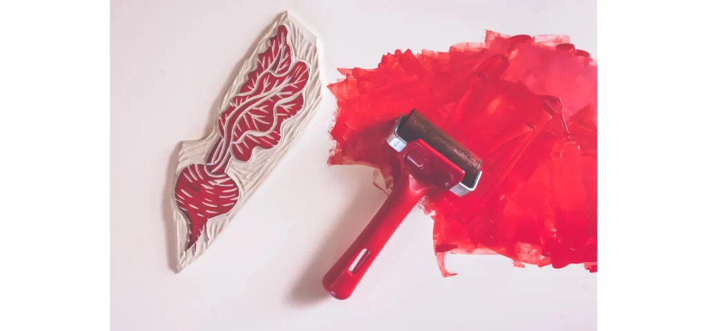 in the center of the image is a red brayer with a black roller. it is sitting on a patch of rolled out red paint. next to it on the left is a stamp with an image of a butterfly on it that has the red ink on it. the details of the stamp are red and the negative space on the stamp is white. they are all on a white background