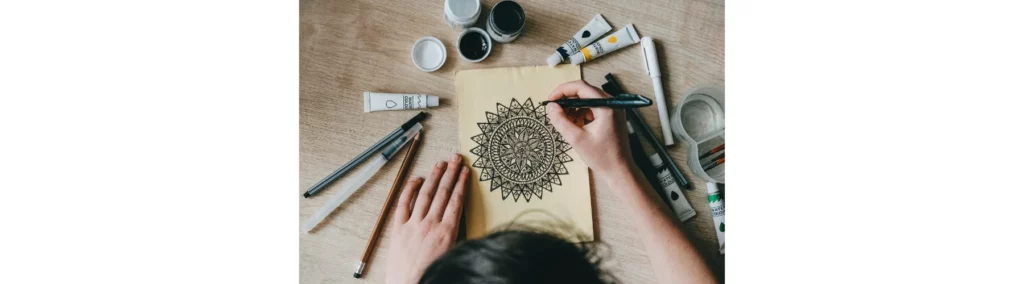 in hte center of the image is a waoman leaning over a table drawing ona beige paper. she is drawing a mandala and its in black ink. around her are various art supplies. on a wooden desk top surface