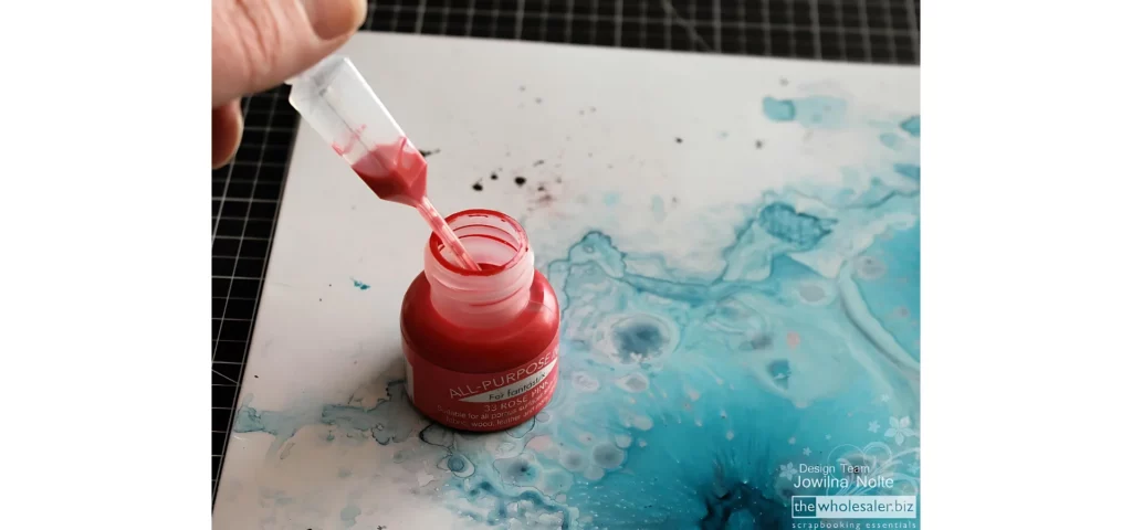 in the center of the image there is a bottle of red ink that is open. there is a piipette pulling ink out of the bottle. it is sitting on paper that has blue ink on it to make it look like an ocean. the top left coener you can see just a small part of a persons hand holding the pipette