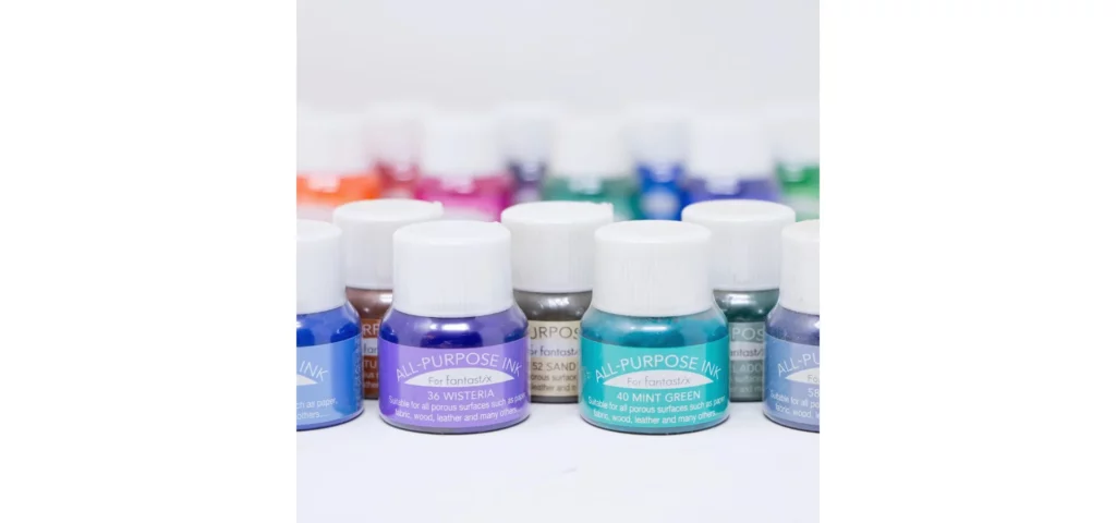 in the center of this image are multiple bottle of different coloured inks all lined up in uneven rows. behind them are more but they are slightly blurred out. on a white background