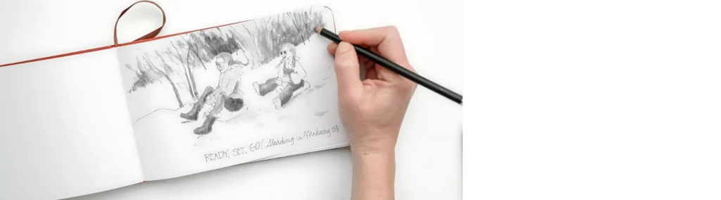 in the center of the image is an open landscape sketchbook that on the right hand side has ha sketch of a couple sitting in a park on it in graphite pencil. there is a hand to the right hand side holding a black cased pencil and sketching the trees. the book has a red cover which is visable and a red ribbon on the top of it. on a white background