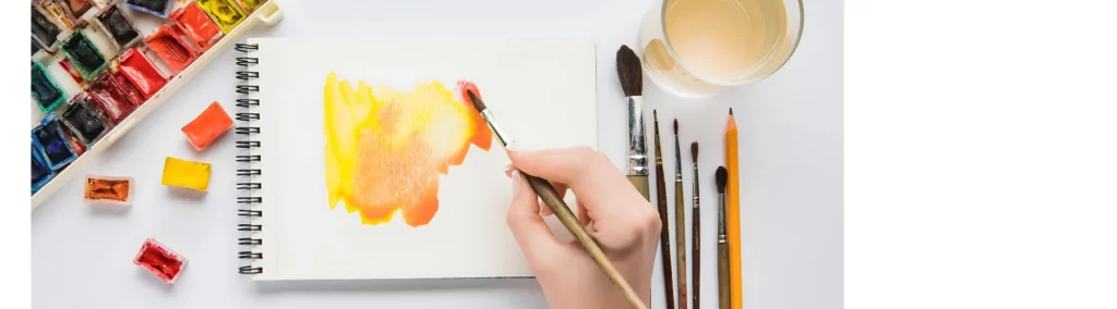 in the center of the image is an open ringbound watercolour pad that is being painted on with orange and yellow watercolour paint using a long stemmed paintbrush. it is very watery. to the right are variuos sizes of brushes and a glass of water. to the left is a set of watercolour pans that have been used in their tin and some loose pans sitting on the white desk