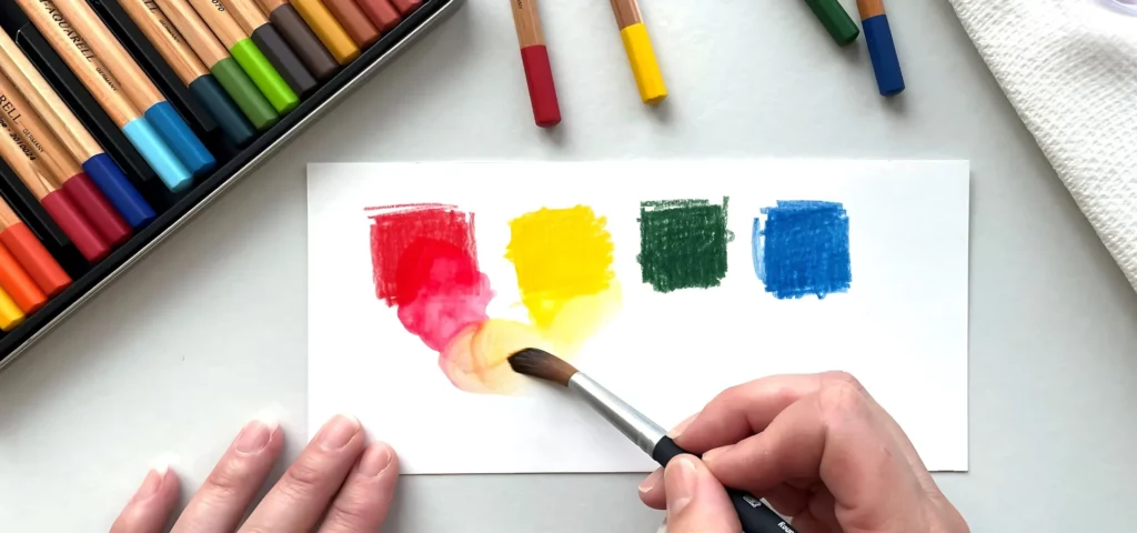 in the center of the image there is a pair of hands. the right hand is holding a paintbrush. the left hand is holding the paper down. the paper has four colour swatches on it. from left to right it is red, yellow, green and blue squares. the paintbrush is blending out the red and yellow colour on the paper. on top of the paper are 4 pencils in the same colour lying on the table. on the left of the paper is a tin of watercolour pencils. you can only see the ends of the pencils showing the colours of them. they are all on a white table background. there is a white paper towel to the right hand side of the image.