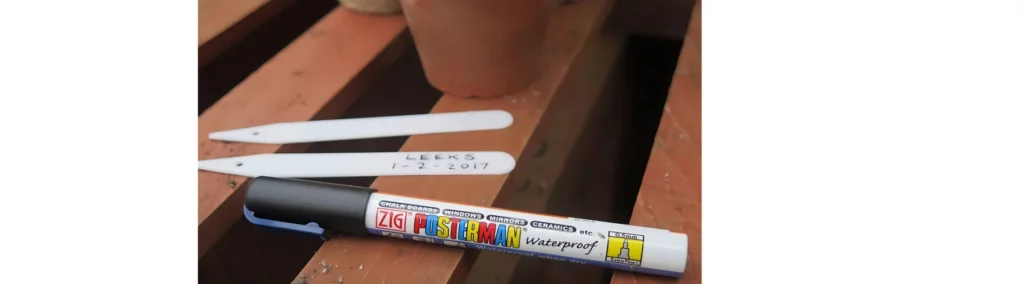 in the front center of the image is a black posterman pen cap on facing the left side of the image. sitting on wooden slats with a terracotta pot behind it. behind it are two plastic plant name sticks with writing on it. showing how the pen works on different surfaces. the picture has been taken outside.