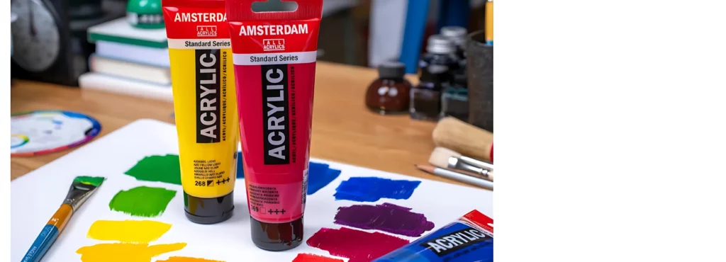 in the center of the image there are two tubes of amsterdam acrylic paint. a red and a yellow that are standing ona  colour chart made out of the paints. there is a paintbrush on the left that has green paint on its hairs and a small colour chart in the background. the background is an art studio with various art items visable.