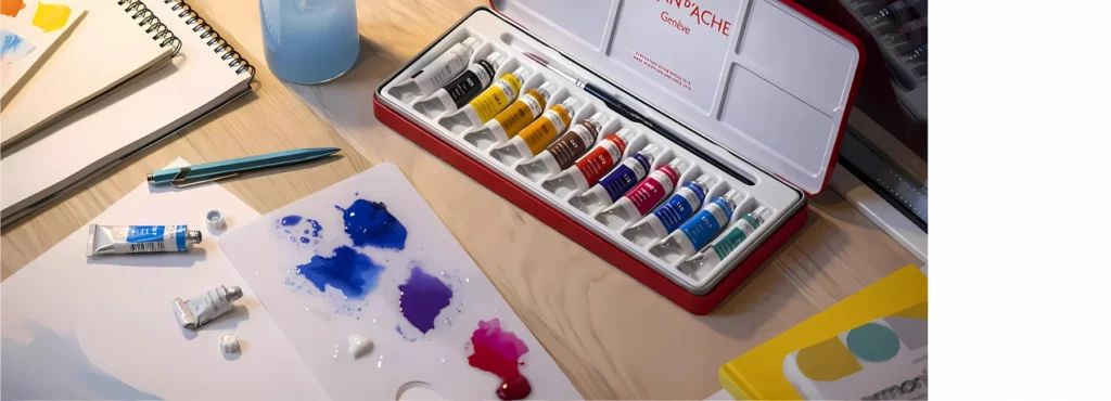 there is a plastic palette with some purple, blue and pink gouche on it mixed with some water. there are tubes of gouache open and lying on some paper infront of that palette. behind it is a set of gouache paints in a tin. to the left are various pads of paper and a glass with blue tinted water in it.