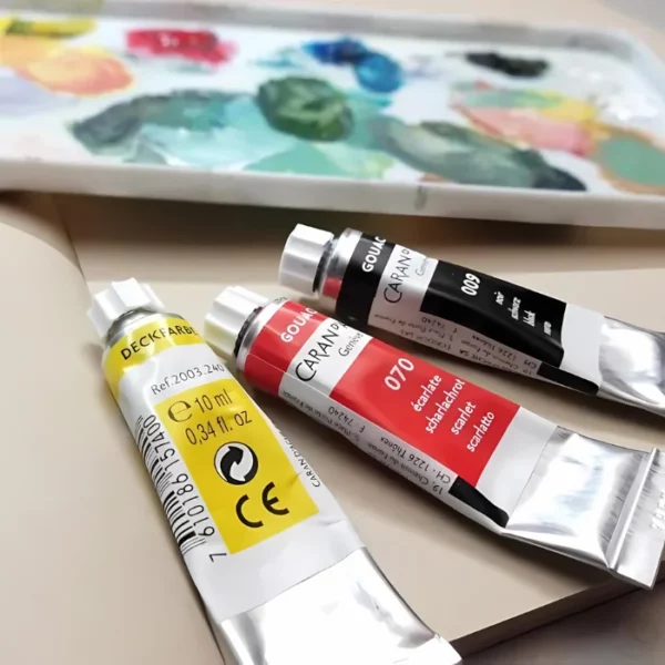 Three tubes of Caran D'Ache Studio Gouache Paint are seen laying on a pad with a plastic pallete covered in ink, in the background, which is slightly blurred.