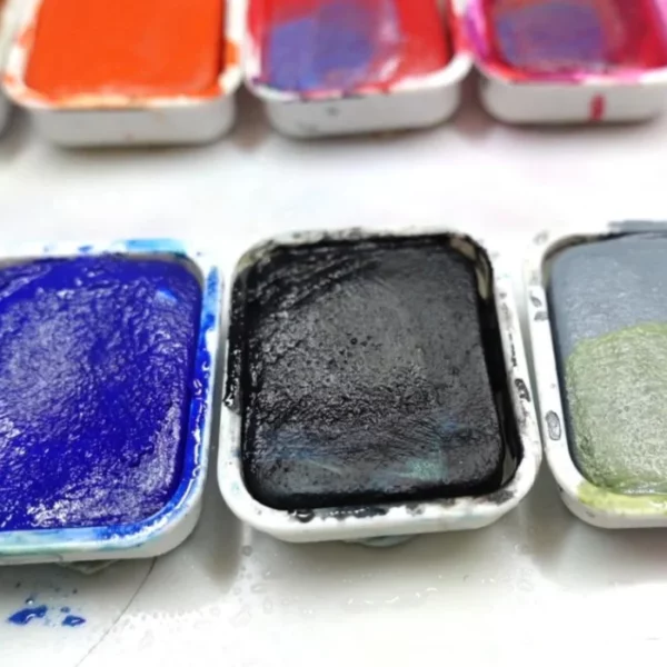 A close up of 3 pans of Caran D'Ache Studio Gouache Cakes. There are three in the background that are slightly blurred. They cakes at the front are blue, black and grey. They have been used. The image is cut off by the frame.