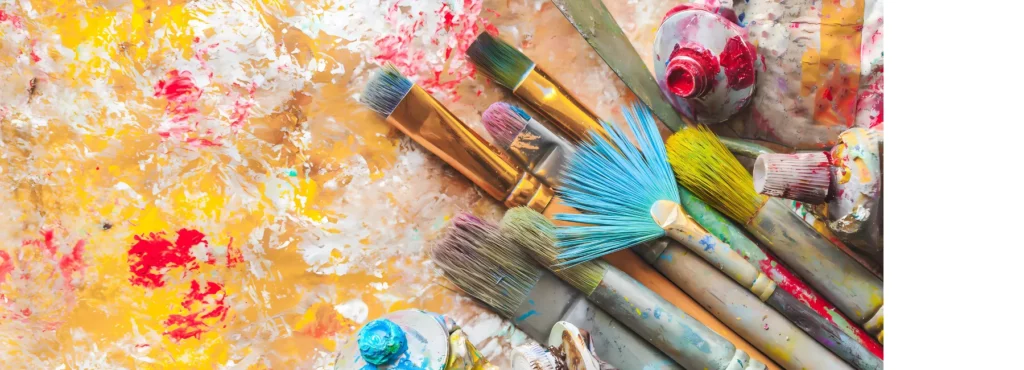 on the right of the image is a collection of different types of brushes and tubes of oil paints. they are lying in at an angle towards the center of the image. they ahve all been used and their hairs are dyed all different colours and the handles have dried paint on them. they are resting on a canvas that has been painted in oil paint in various colours and has texture to it