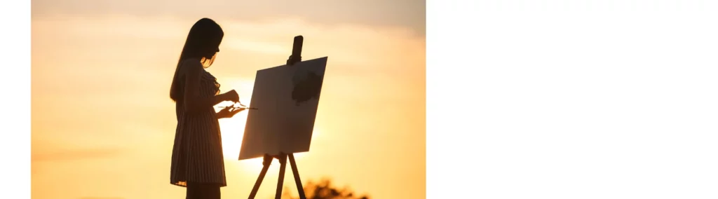 there is a slight silhoutte of a lady in a sundress standing on a hill with the sun setting behind her painting on a standing easel. she has long hait. the picture has yellow tones to it.