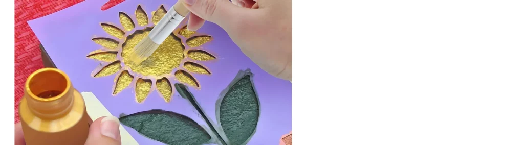 in the center of the image there is a stencil of a sunflower with green leaves and yellow flower at the top. someone is using a stippling brush to paint the flower through the stencil. a tub of yellow paint is open on the left hand side of the image