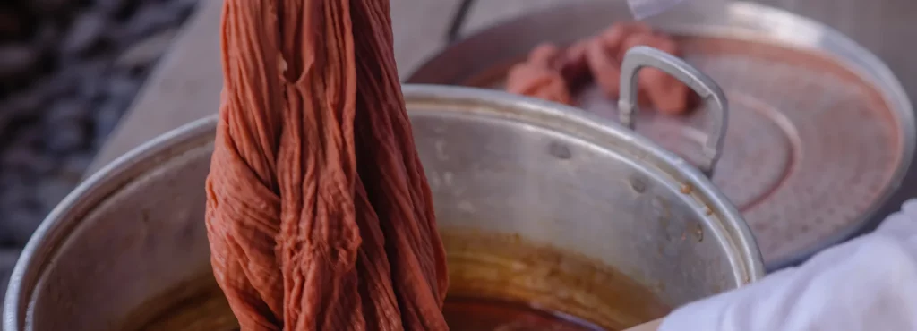 in the center of the image is a large metal silver pot that has orange fabric dye inside of it. a shirt is being pulled out of the mixture with the dye on it. in the background you can see the lid of the pot resting on a table top