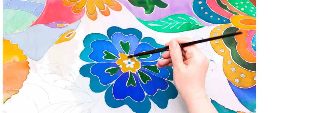 in the center of the image there is a fabric with various flowers and leaves drawn and painted onto it in different colours. there is someone painting onto the fabric with a paintbrush.