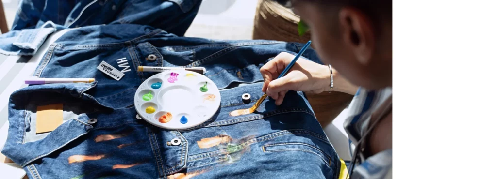 in the center of the image is a denim jacket that is laid out on the table and someone is sitting to the left of it painting onto it with a brush with the orange colour stripes. there is a palette in the centre and the palette has variuos different colours of fabric paint blobbed into it.