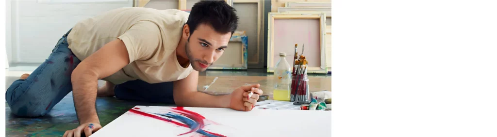 the image is of a canvas that has been gessod. there is aman kneeling on the floor over tha canvas and painting swirls in bright colours of red and blue. he is concentrating on the canvas and has his bottle of brushes next to him. he has a wall behind him with various sized canvas leaning against it