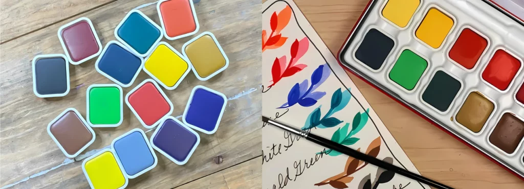 on the left are a bunch of loose gouach cakes scattered on a wooden table, they are a variety of different colours. on the right is a set of the gousche cakes and they are next to a paper that has the names of the colours and a colour sample next to them in a shape of a leaf. showing how they look wet and dry. on a light wooden table top