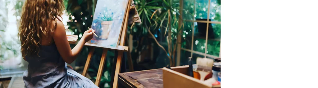 the image is of a woman with long brown hair sitting infront of an easel with a canvas on painting pictures of potted plants. the background is her painting room and has various are supplies areound but also lots of plants around it. the weather can be seen and it is rainy