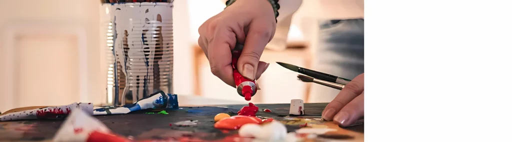 in the center of the image is a person holding and squeezing a tube of red oil paint onto a wooden palette. there are other paints already on it. on the left is a tin with white paint that has run down the side of it. they are holding two paintbrushes in their other hand