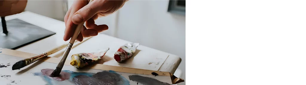 in the center of the image there is aperson using a paintbrush on a mixing tray mixing paint and mediums together. there are various tubes of paint that are open and paintbrushes are laying around everywhere. it is on a table surface and the background is a blurred visual of the room