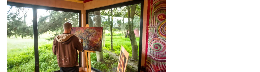 in the center of the image is a man in a hoodie standing infront of an easel painting an abstract picture. he is in a glass room and you can see his garden through the windows. it is daytimes and misty and bright outside