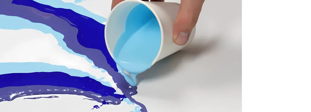 on the left side of the image there are three shades of blue and a white paint that have been poured onto the white canvas ina triangle type shape. there is a white cup at the tip holding the paint that is being poured out. by a person holding it. you can see their thumb and some of their hand