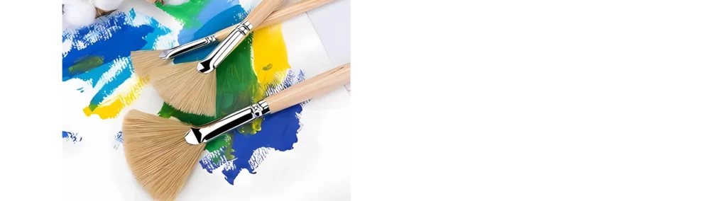 in the center of the image there are three different sized fan brushes lying next to and on top of eachother. they are resting on a canvas that has a mix of blue, yellow and green paint on it.