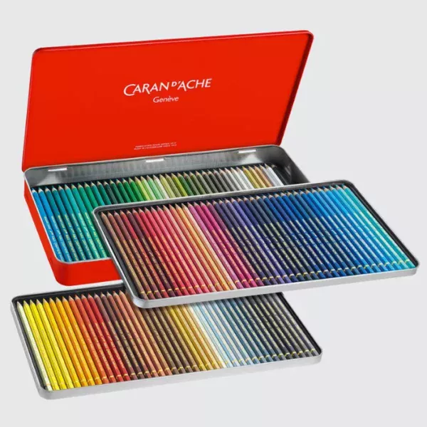 A Set of 120 Caran D'Ache Pablo Coloured Pencils is shown in the center of the frame. The set is opened and there are 3 trays of pencils that are shown coming out of the red tin. The tin lid is open and the Caran D'Ache logo is printed inside the tin in white. The pencils are lined up next to each other in each tray. They have a coloured barrel that depicts the colour of the pencil. On a light grey background.