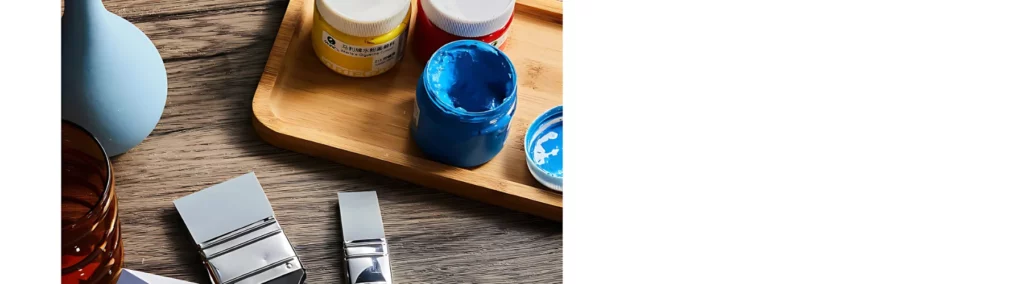 at the front of the image are two silicone brushes resting on a wooden table. they are next to a brown tray that has three pots of thick paint on it. the blue paint j=has its cap off. on the left is a glass jar and a pot.