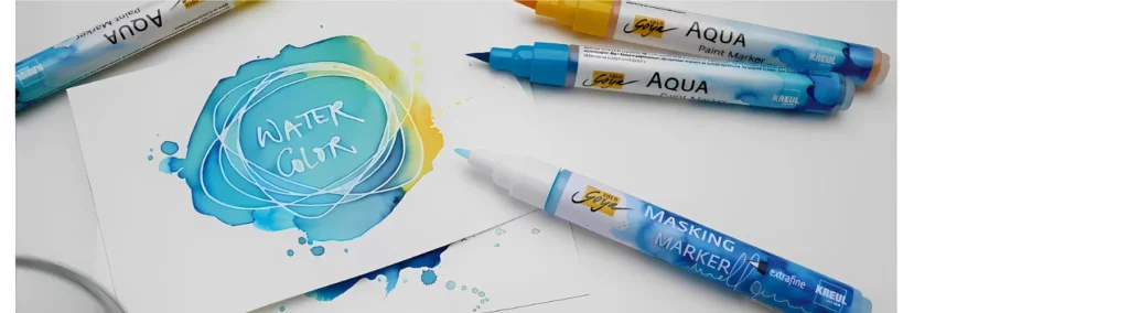 in the center of the image is a watercolour card that has been painted in blue and yellow. it has watercolour blocked out of it in the centre. on top of it are various pens and the solo goya masking markers are lying on the table.