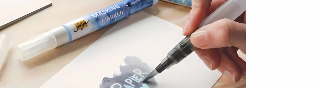 at the front of the image is a small paper that has writing on it and watercolour in a dark gray colour. someone is painting over it. they have used the masking fluid to mark out a word in the centre and are now painting over it. behind the hand is a masking pen lying on the table