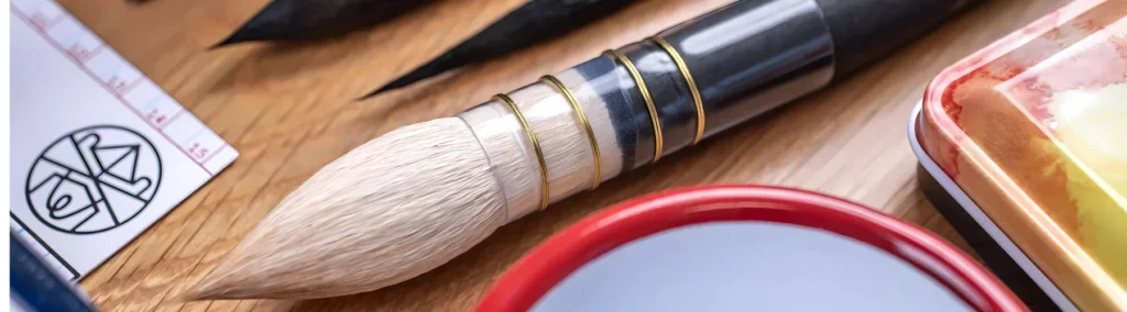 in the center of the image is a close up of a tintoretto brush. you can see it has a black handle and a white rounded haor. next to it you can see the tips of two black haired brushes. and a watercolour pan set on the right with a bowl next to it. they are all lying on a wooden table