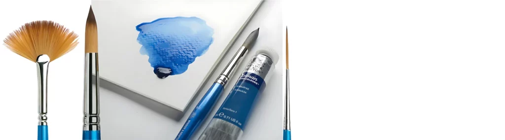 on the left of the image is a fan brush with a blue handle and silver ferrule and orange hair. next to it is a round brush with a blue handle silver ferruler nad orange hair. in the center is a pad of paper with blue watercolour paint in the corner that is pooled, next to a brush and a tube of winsor and newton watercolour paint. on the right of the image is a liner brish that has a blue handle with a silver ferrule and orange haor. on a white background