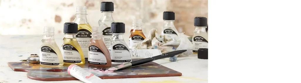 there are various bottles of winsor and newton oil mediums spread out across the image. they are all clear bottles with black and white labels and black lids. they are various sizes and all different products. they are resting on a wooden palette with a paintbrush and a tube of oil paint resting on it. the background is a blurred visusal of the room