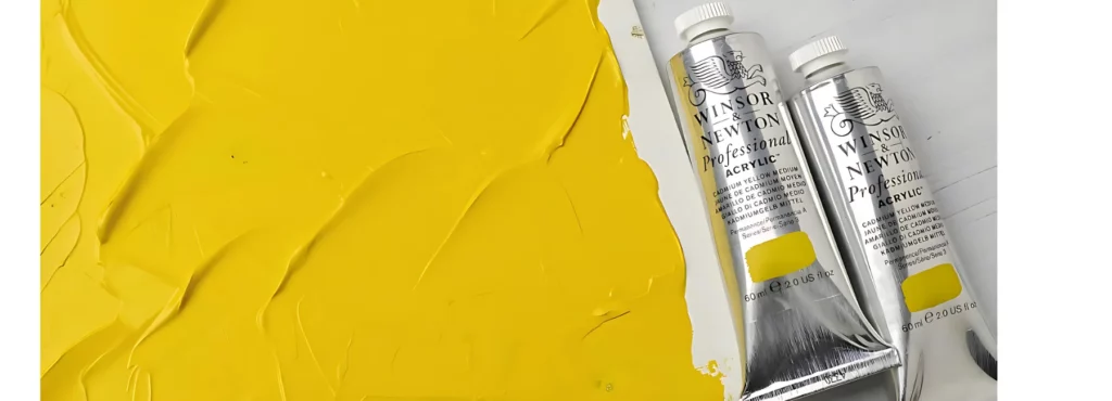 the left of the image is a canvas panel that has been thickly covered in yellow acrylic paint. to the right are two tubes of proffessional acrylic paint in their silver tubes with the white caps on a white table top