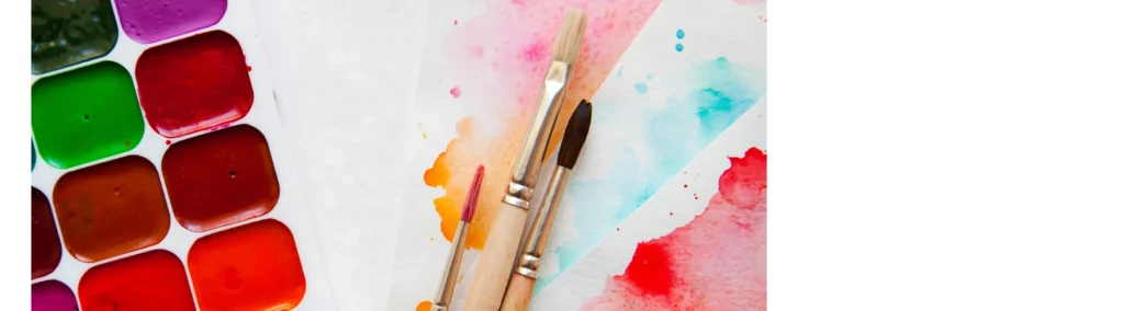 on the left hand side is a palette with various watercolour paints inside of it. on the right are three brushes lying on watercolour paper that has watercolour painted onto it