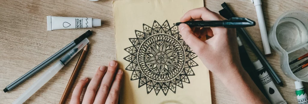 the image is a birds eye view of someone drawing a mandala pattern onto brown paper using black fineliners. around them are various pens and paints. on a brown tablke