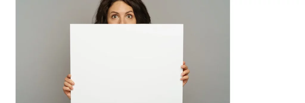 the image is of a woman holding up and standing behind a large white board. you can just see her fingers holding the board and her eyes and head above it. she is standing infront of a grey wall