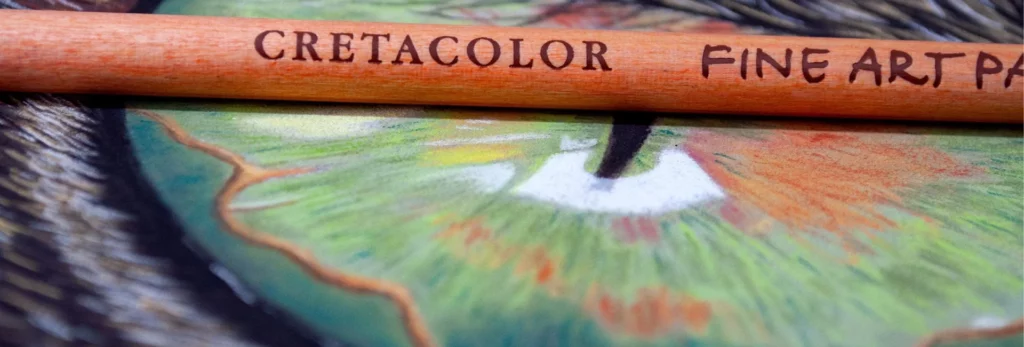 the image is a close up of a  single pastel pencil. it is lying on top of the image and you can just see the name and what pencil it is written on it. beneath it is an image of an eye that has been drawn by the pencils