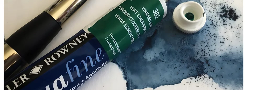 the image is a close up of a single tube of aquafine watercoour paint. in viridian green. the tube has a white cap which is off and there is paint beneath it on watercolour paper. the paint is granulating slightly. there is a watercolour paintbrush to the left of the tube of paint.