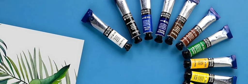 the image is a birds eye view of a selection of tubes of the aquafine paint in a half moon shape. they all have black caps and are in rainbow order on a blue board. to the left is a watercolour paper with an image of a plant painted on it using the aquafine paints.