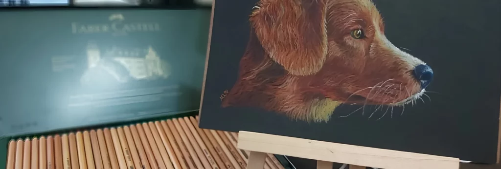 the image is showing an open tin of pastel pencils and you can see the faber castell branding on the tin. to the right of the tin is a sketch of a brown dog on black pastel paper sitting on an easel.