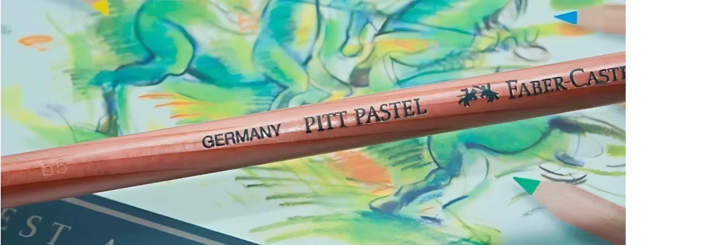 the image is a close up of a single pastel pencil infront of a tin of pastel pencils. the pencil is encased in wood and the name and brand is clearly visable on it.