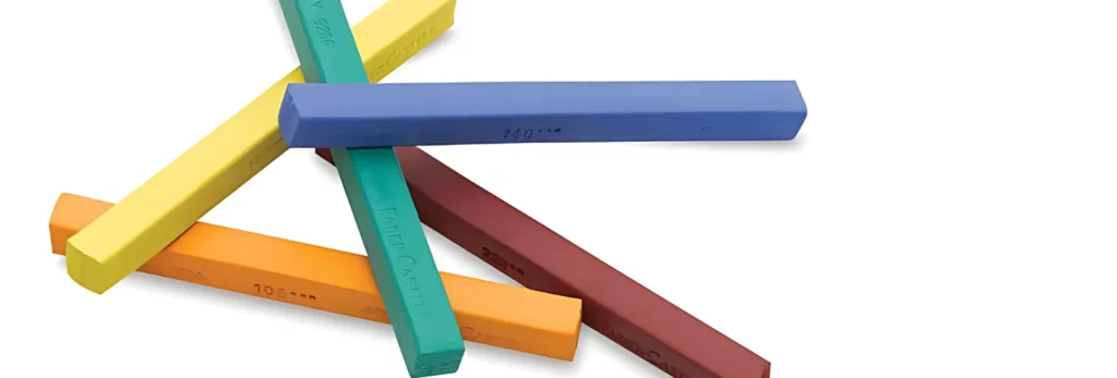 the image shows a close up of five pastel sticks all haphazardly stacked on eachother. they are on a white background. the colours of the sticks are: yellow, orange, green, red and blue. they are long and thin and swuare in shape.