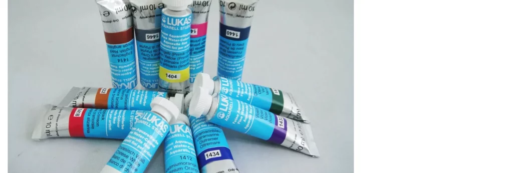 the image is a close up of a stack of lukas watercolour paints. they are in the center of the image and have blue paper covered tubes with the names and colours of the paint on it. they have white caps and are stacked ontop of eachother in the front and in the background they are standing on their caps. on a white background