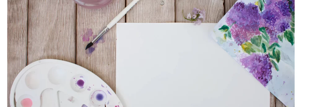 the image is a birds eye view of an area being used for watercolour painting. there is a palette on the left with paint in it. a brush at the top of the image leaning on the table and on the paper and to the right is a painting of flowers done in the paint. all the paints are shades of purple and pink. the table is a light wooden table