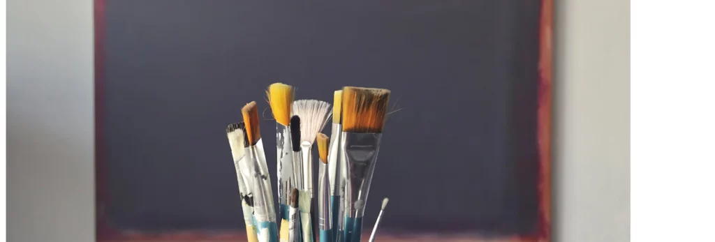 the image is of a close up of a group of paintbrushes in a jar and behind it is a primed black canvas ready for painting. the background is a blurred image of a room