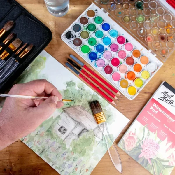 A close up of a persons hand holding a brush from the Mont Marte Acrylic Handle Watercolour Brush Set and painting a picture of a house on watercolour paper. There is a paint palette and some brushes scattered around the painting.